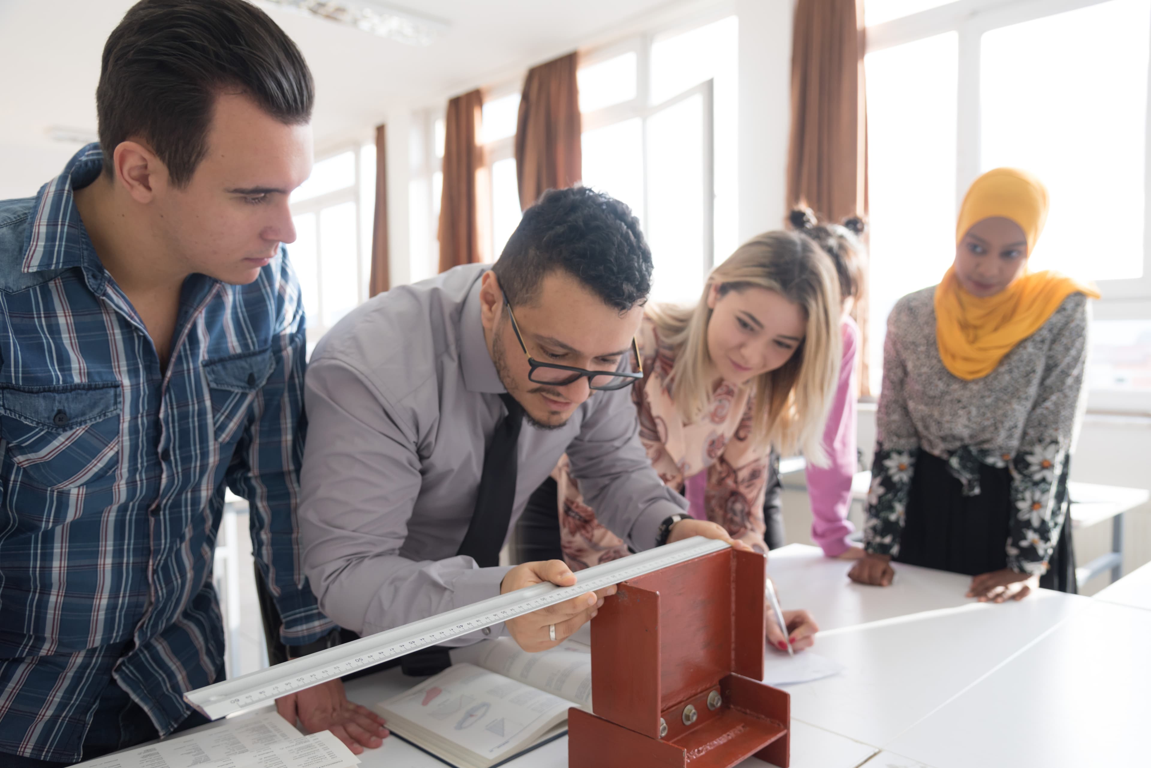 A diverse group of students at a technical college work together around a table, taking notes and measuring a red metal structural I-beam model during a civil engineering or construction class.
